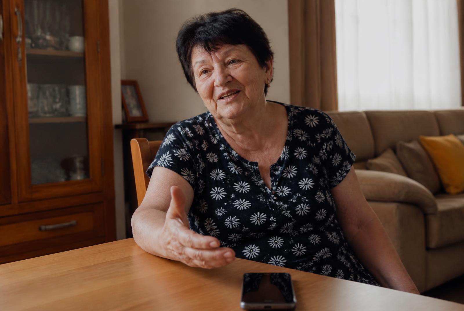Older woman sitting at a table at home telling a story aloud toward a smartphone